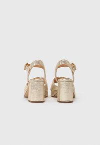 Gyldne, metalliske hælede sandaler med en kraftig blokhæl, ankelrem og åben rygdesign. Struktureret overflade med synlige syninger.