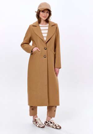 Manteau long de couleur camel avec un col et deux boutons, associé à un haut à rayures et un pantalon à accents marron. Les chaussures présentent un motif de vache.