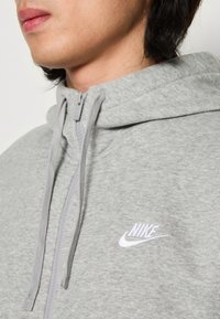 Gros plan d'une personne portant un sweat à capuche Nike gris clair avec une fermeture éclair et des cordons, montrant le logo Nike sur la poitrine.