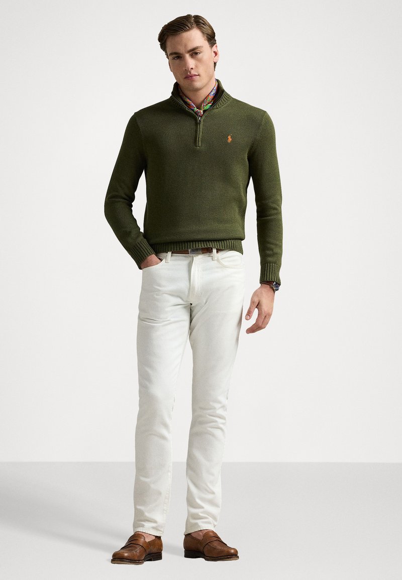 Maglione verde a maglia con mezza cerniera, caratterizzato da un piccolo logo. Abbinato a pantaloni bianchi e mocassini marroni. Visibile il tessuto testurizzato.