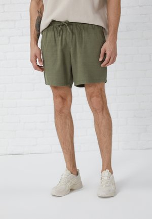 Mann trägt olivgrüne Shorts mit Kordelzug und weiße Sneakers und steht an einer weißen Ziegelwand.