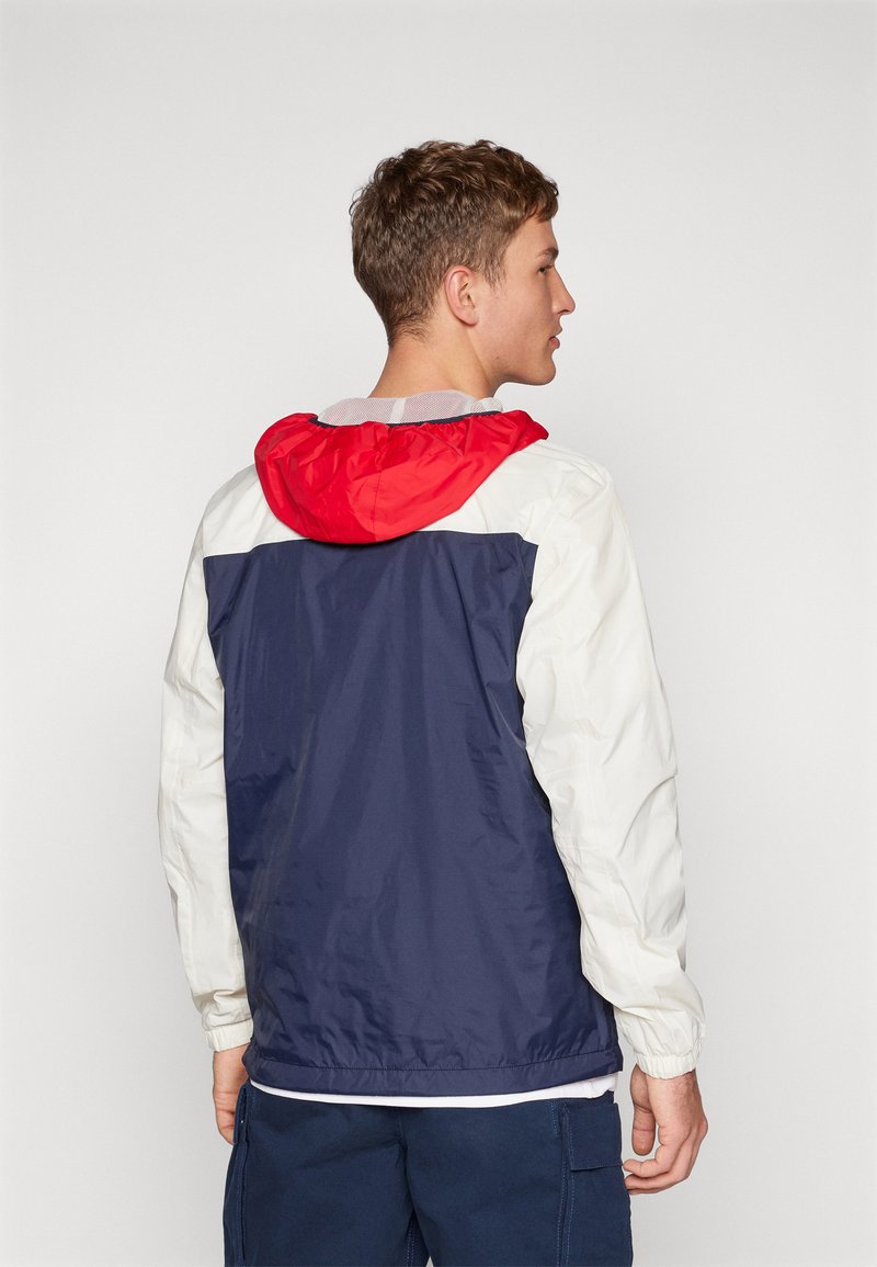 Giacca leggera a blocchi di colore con cappuccio rosso, retro blu e maniche bianche. Polsini elastici e vestibilità rilassata. Il materiale sembra liscio.