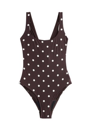 Junge Frau trägt einen dunkelbraunen Badeanzug mit weißen Punkten und steht am felsigen Strand, wobei sie eine Hand auf einen großen Felsen legt.