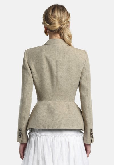 Mujer con coleta torcida rubia que lleva una americana ajustada beige y una falda blanca con volantes, vista desde atrás sobre un fondo blanco.