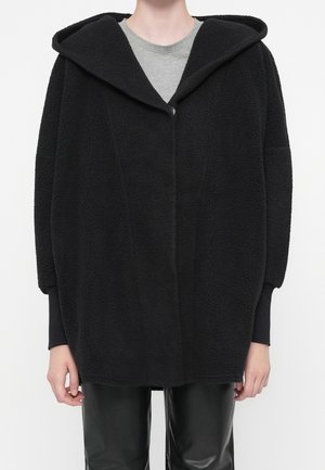 Personne portant une veste à capuche en polaire noire oversize sur une chemise grise et un pantalon en cuir noir, debout devant un fond blanc.