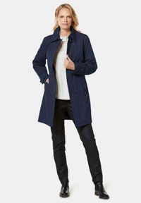 Navy blue trench coat with a collar, double-breasted buttons, front pockets, and a knee-length cut, worn over a light-colored shirt.