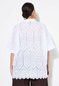 Blusa bianca di cotone con motivi a taglio, maniche corte e orlo scallop. La schiena è decorata con ricami intricati.