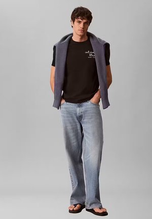 Giovane uomo che indossa una t-shirt nera Calvin Klein, jeans azzurri chiari, infradito, con un maglione grigio drappeggiato sulle spalle, in piedi davanti a uno sfondo neutro.