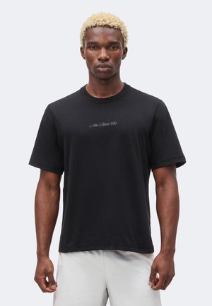 Młody mężczyzna z krótkimi blond włosami, ubrany w czarną koszulkę "Nike Athletic Club" i jasne spodenki, stoi na jednolitym tle.