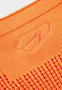 Țesătură tricotată de un portocaliu strălucitor, având un model texturat și un logo brodat în formă de frunză. Designul include zone cu tricot deschis pentru ventilație.