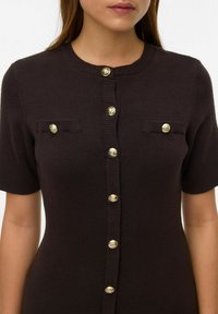 Cardigan marrone scuro in maglia con maniche corte, caratterizzato da bottoni dorati e due tasche sul petto con dettagli a coste.