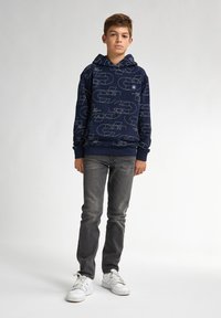 Sudadera marina con patrones geométricos blancos, hecha de una tela suave. Combinada con jeans grises ajustados y zapatillas blancas, de pie contra un fondo claro.