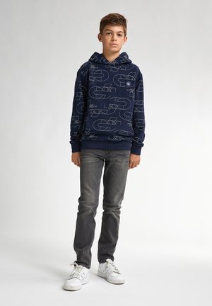Felpa navy con pattern geometrici bianchi, realizzata in un tessuto morbido. Abbinata a jeans grigi slim e scarpe da ginnastica bianche, in piedi davanti a uno sfondo chiaro.
