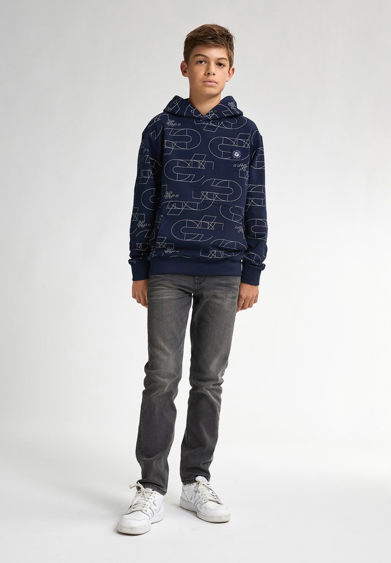 Sudadera marina con patrones geométricos blancos, hecha de una tela suave. Combinada con jeans grises ajustados y zapatillas blancas, de pie contra un fondo claro.