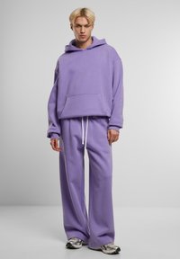 Fioletowy sweatshirt z kapturem i kieszenią z przodu, w połączeniu z szerokimi spodniami dresowymi w tym samym kolorze oraz białymi sznurkami. Nosi się z szarymi butami sportowymi.