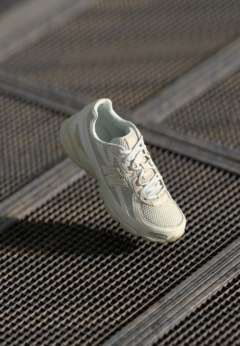 Chaussure de course beige et blanche avec un design en mesh et des lacets reposant sur une surface en grille métallique avec des ombres en angle.