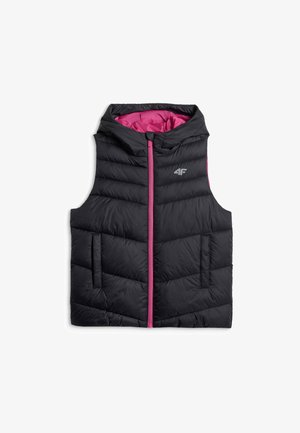 Jeune fille portant un survêtement rose et un gilet matelassé noir, debout devant un fond gris uni.