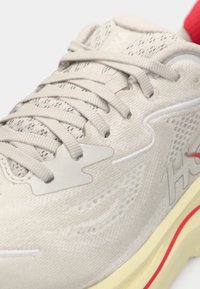 Scarpa sportiva grigia chiaro con tomaia in rete, lacci piatti, dettagli texture, linguetta rossa sul tallone e intersuola gialla con dettagli bianchi.