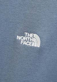 Μπλε ύφασμα με άσπρο κεντημένο λογότυπο The North Face που περιλαμβάνει κείμενο και σχέδιο ημικύκλου.