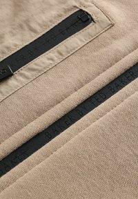 Beige kangas, millel on tekstuurne pind ja mustad detailid. Omab silmapaistvat logosilt, millel on kiri "BAKER BY TED BAKER" ning lukuga taskut.