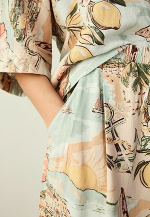 Hand steckt in der Tasche eines pastellfarbenen Outfits mit nautischen und Fruchtmustern auf weichem Stoff.