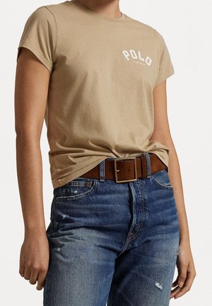 Personne portant un t-shirt beige "Polo" rentré dans un jean bleu déchiré avec une ceinture en cuir marron avec une boucle carrée dorée.