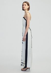 Weißer Jumpsuit mit schwarzen Seitenpanelen, botanischem Druck und verstellbaren schwarz-weißen geflochtenen Trägern. Langer, lockerer Schnitt mit einer Schleife.