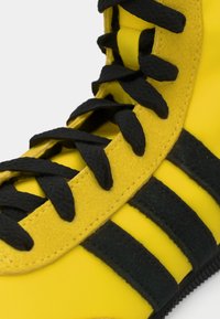 Yellow athletic shoe with black laces and three black stripes. Textured materials highlight the design and color contrast. Flexible sole.