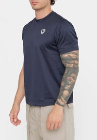 T-shirt blu navy in tessuto morbido, maniche corte, colletto rotondo sottile, con un piccolo logo a scudo sul petto, abbinato a pantaloni beige.