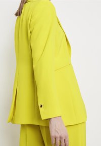 Blazer giallo brillante con una vestibilità su misura, tessuto liscio e dettagli con bottoni dorati sulle maniche. Presenta una fessura laterale per facilitare i movimenti.