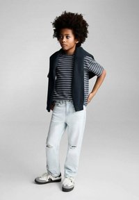 Otrok nosi prugasto sivo-belo majico s temnim pletenim jopičem, svetlo modrimi raztrganimi kavbojkami in sivimi supergami z belimi vezalkami.