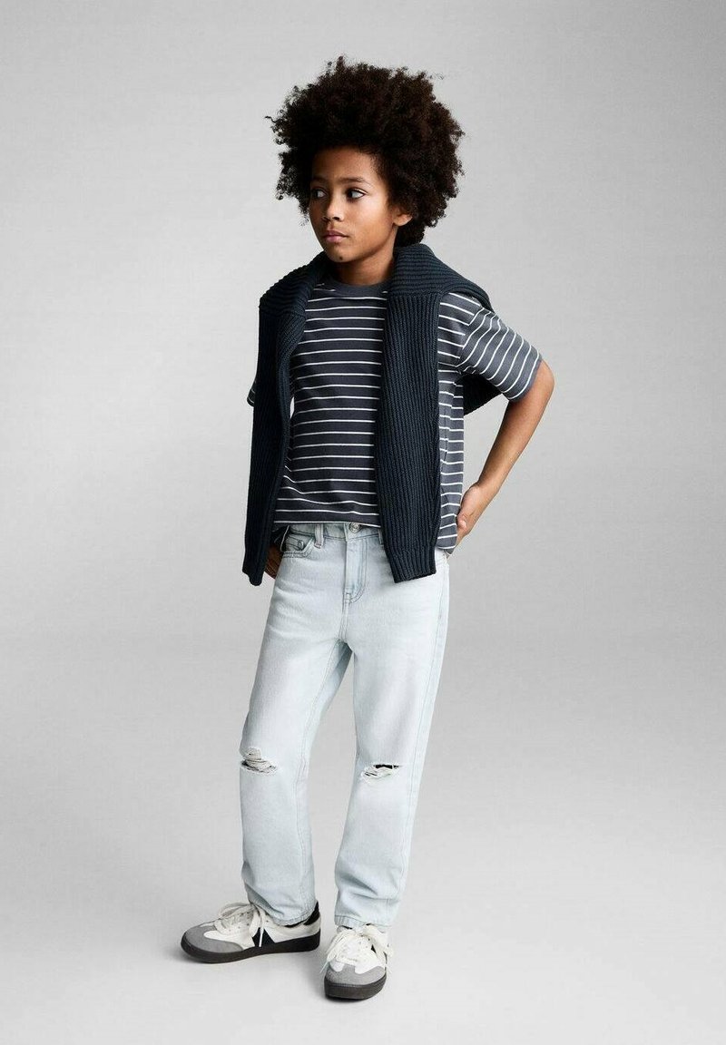 Otrok nosi prugasto sivo-belo majico s temnim pletenim jopičem, svetlo modrimi raztrganimi kavbojkami in sivimi supergami z belimi vezalkami.