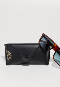 Lunettes de soleil Ray-Ban en écaille de tortue brun avec des verres teintés verts reposant sur un étui Ray-Ban en cuir noir texturé.