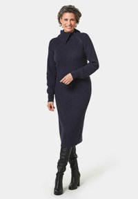 Strickkleid in Dunkelnacht mit hohem Kragen und Reißverschluss-Details. Figurbetont, lange Ärmel und eintexturiertes Rippdesign. Knielang.