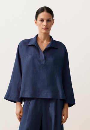 Woman with dark hair in a low bun wearing a loose navy blue long-sleeve shirt with a collar and matching wide-leg pants.