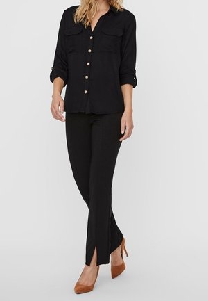 Frau trägt eine schwarze Bluse mit Knopfleiste und hochgekrempelten Ärmeln, schwarze schmale Hose mit vorderen Schlitzen und braune Wildleder-Pumps.