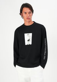 Zwarte lange mouwen t-shirt van katoen, met een grafische afdruk van een model op de voorkant en witte tekst langs de linkermouw. De stof heeft een gladde textuur.