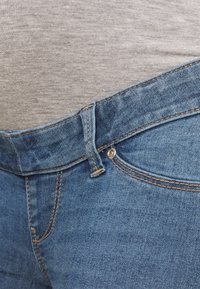 Jeans aus Denim in Blautönen mit sichtbaren Nähten, ausgestattet mit einem Knopfverschluss und Taschendetails. Kombiniert mit einem hellgrauen Hemd.