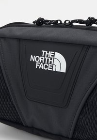 Pochette en tissu noir avec fermeture éclair en haut, présentant le logo "The North Face" en blanc sur un patch en caoutchouc centré sur le panneau avant.