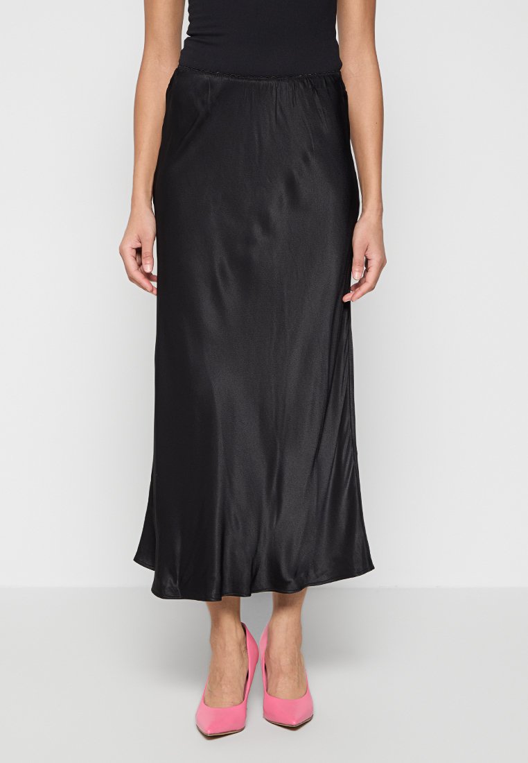 Frau in einem schwarzen Satin-Midirock und leuchtend pinken High Heels, die vor einem schlichten weißen Hintergrund steht.