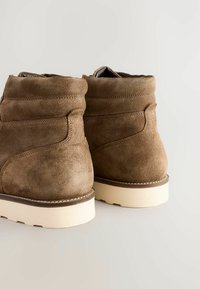 Botas ankle de ante marrón con punta redondeada, textura suave y suela de goma blanca. Presenta costuras visibles y un cuello ligeramente acolchado.