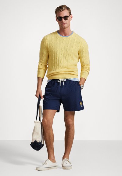 Geltonas megztinis su rialais, tamsiai mėlynos maudymosi šortai su siuvinėtu raštu, balti sportbačiai ir saulės akiniai, laikantis kremiškai baltos ir tamsiai mėlynos rankinės.