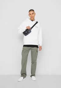 Hvid hoodie over en sort skjorte, olivengrøn cargo-bukser, holder en sort taske med blomstermønster og hvide sneakers. Enkel baggrund.