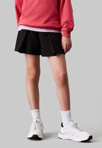 Felpa rossa, gonna nera a pieghe, scarpe bianche con dettagli neri. Calzini bianchi visibili sopra la linea delle scarpe. Tessuto liscio e vestibilità casual.