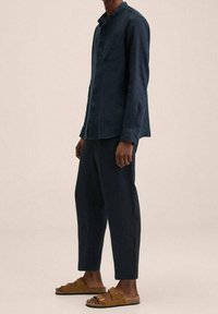 Camicia button-up in lino blu scuro a maniche lunghe abbinata a pantaloni tapered coordinati e sandali in suede marroni con due cinturini regolabili.