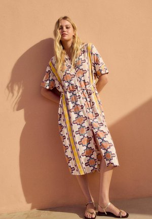Abito camicia con maniche corte, motivi astratti arancioni e blu su uno sfondo chiaro, legato in vita, lunghezza al ginocchio, indossato con sandali beige.
