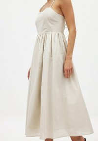 Robe en lin beige avec un corsage ajusté et une jupe évasée, dotée de fines bretelles et d'un tissu texturé, tombant sous les genoux.