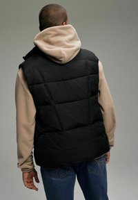 Czarna kamizelka puchowa na beżowym polarowym hoodie, z pikowanym wykończeniem i warstwowym designem, w połączeniu z niebieskimi dżinsami.