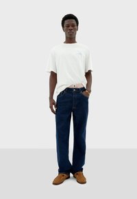 Weißes T-Shirt mit kurzen Ärmeln und blauem Design, kombiniert mit dunkelblauen geraden Jeans und braunen Slippern. Einfaches, lässiges Outfit.