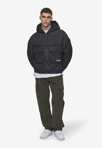 Giacca nera imbottita con cappuccio e zip a metà, due tasche frontali e orlo elasticizzato; indossata sopra una camicia bianca e abbinata a pantaloni cargo verde oliva e sneakers bianche.